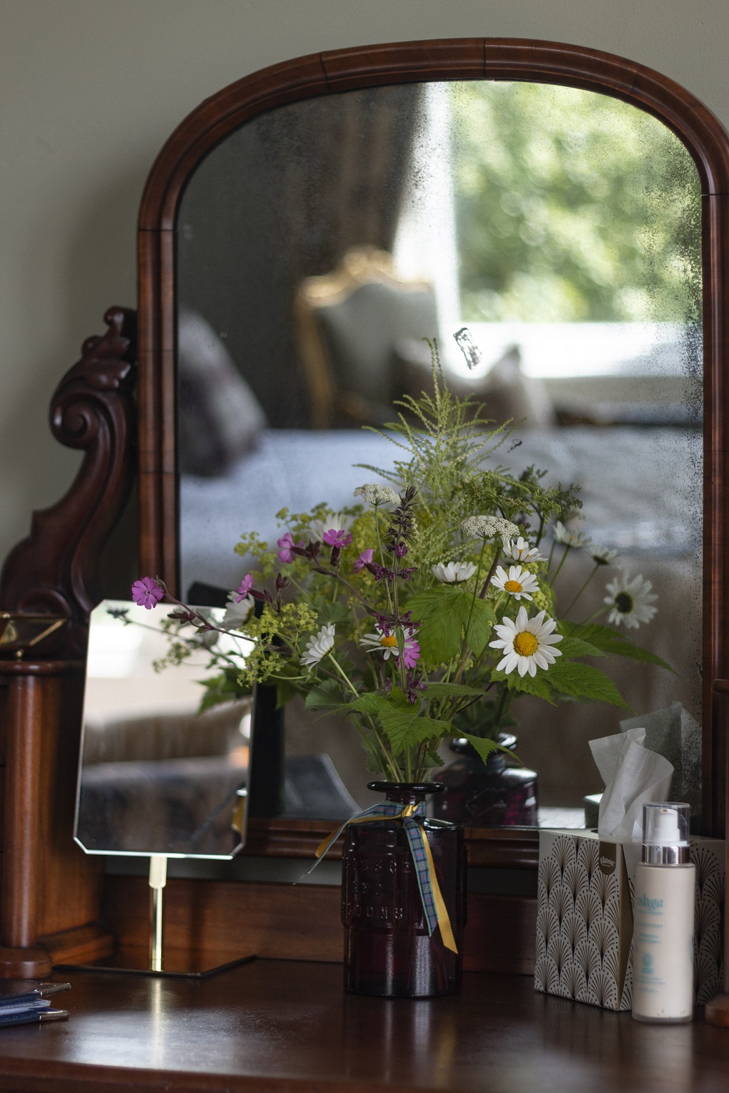 A mirror in a bedroom with a vase of flowers in front of it.