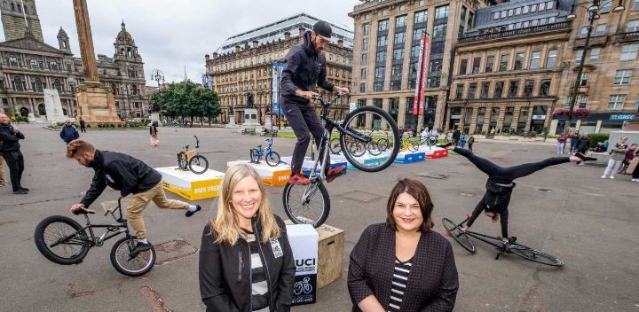 Two women pose for a photo in front of people doing stunts on bikes in a city centre