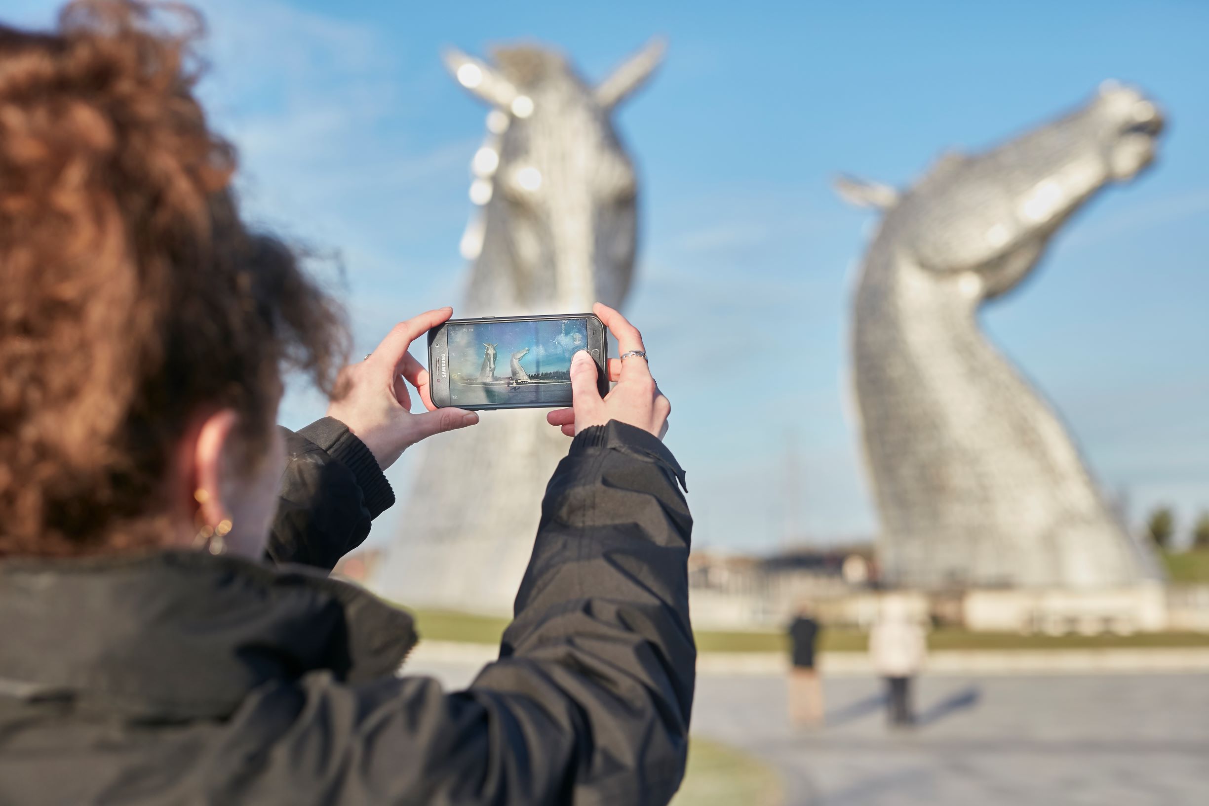 A lady holds a smartphone up to take a photo to two large equine sculptures
