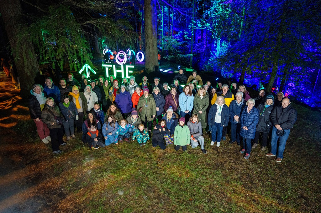 a large group of people standing outside at night, in a forest lit up with sound and light