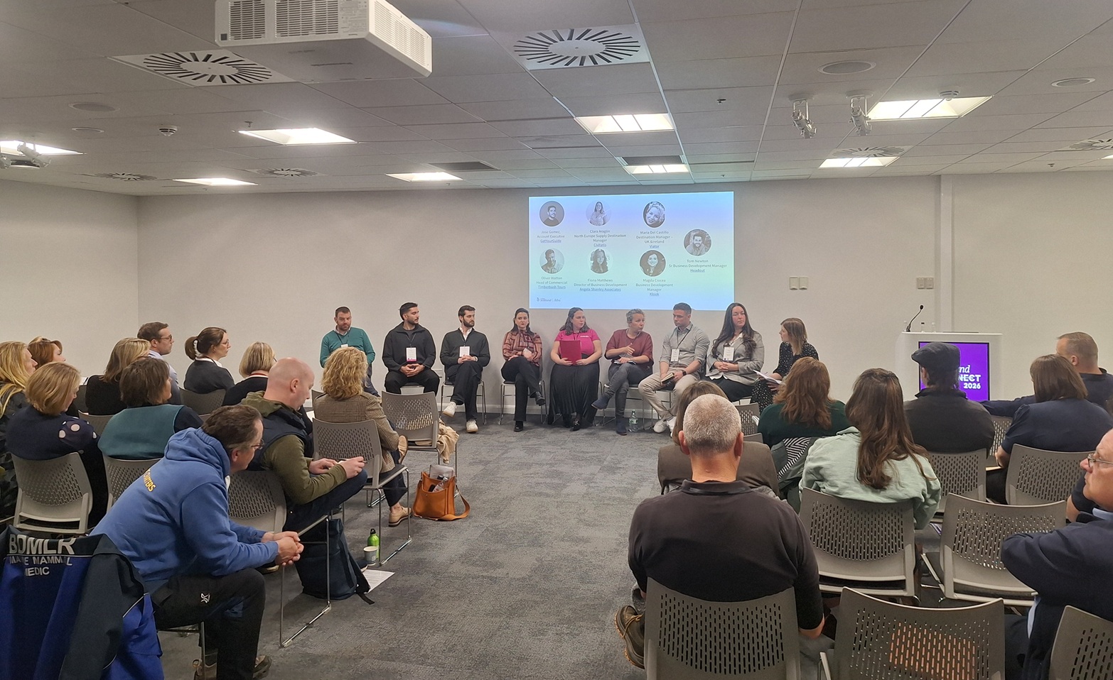 a threatre styke set up of attendess listenogn to a panel session of speakers in a event space