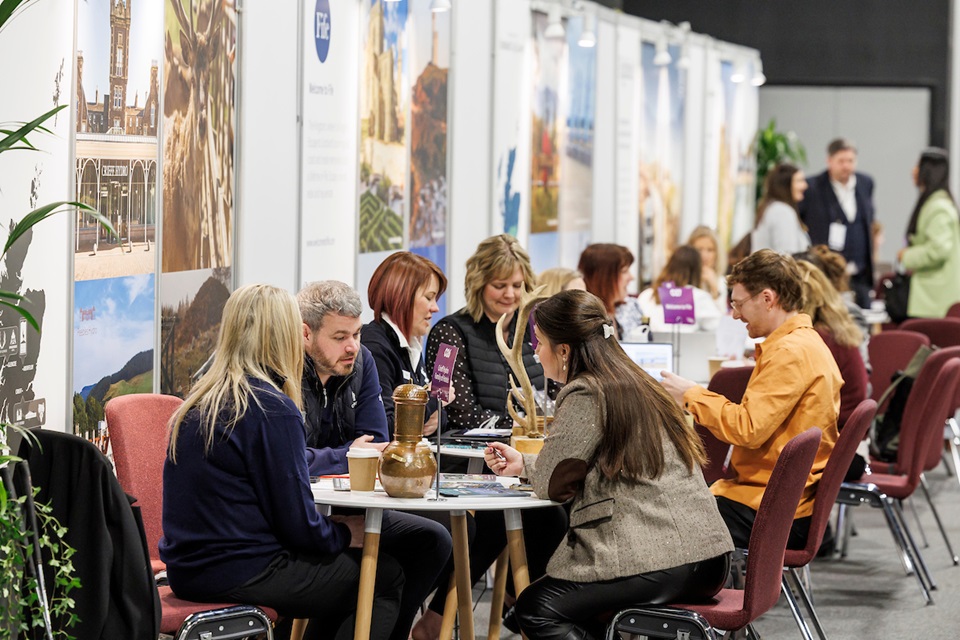 A large group of people sitting having smaller meetings in a large venue with a backdrop of colourful destination images on banners