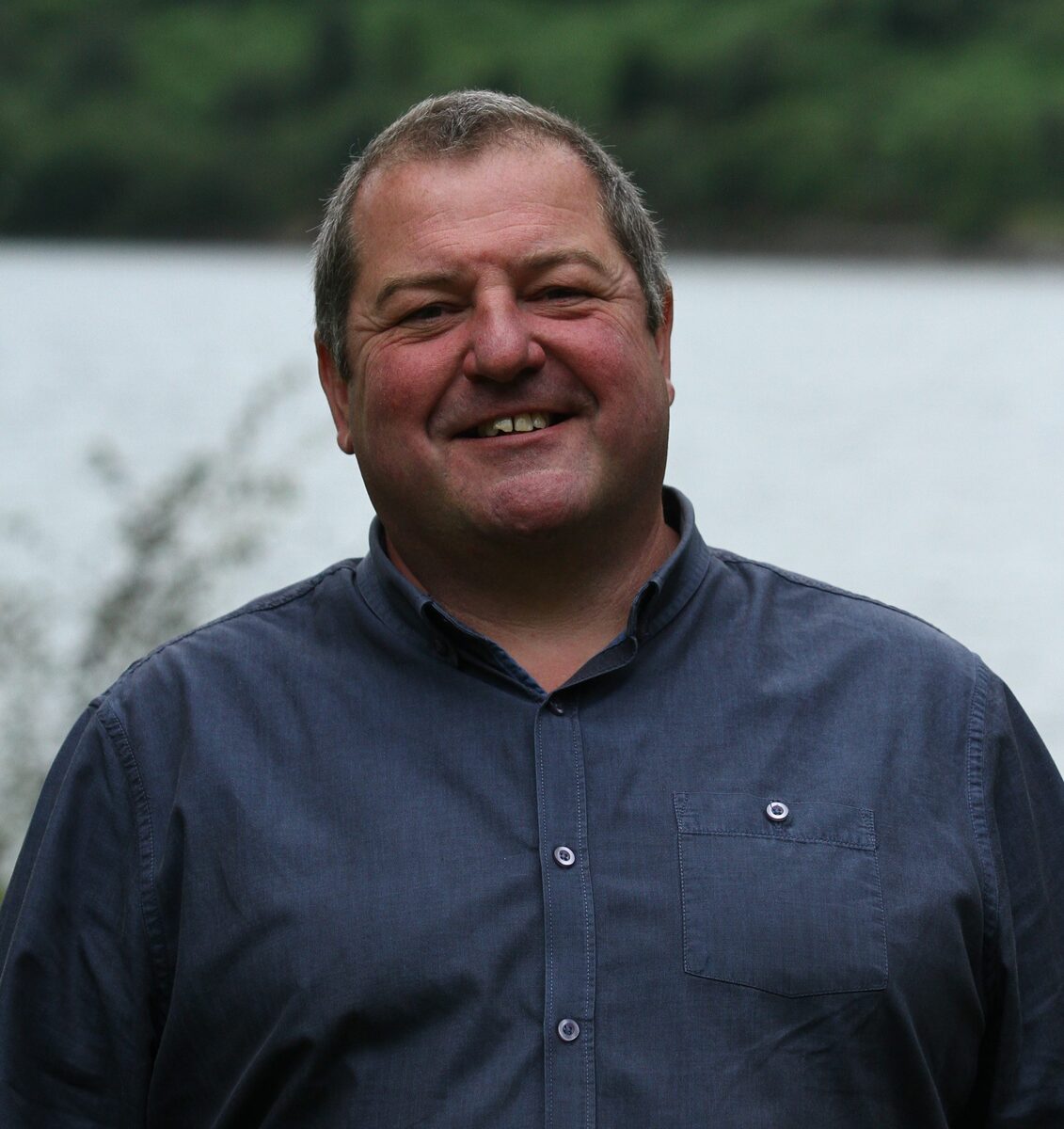 Image of a man smiling to the camera. There is a loch in the background.