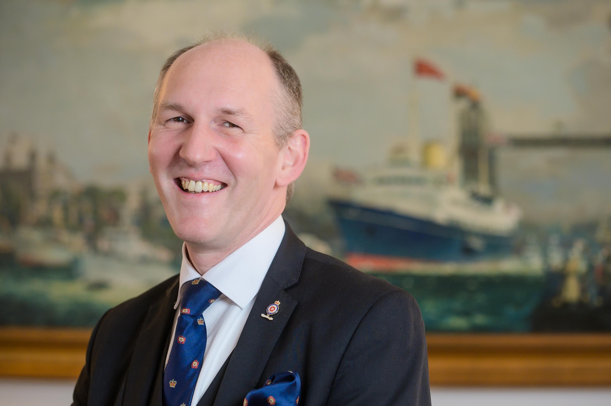 a person smiling, and standing infront of a painting of a ship in the background
