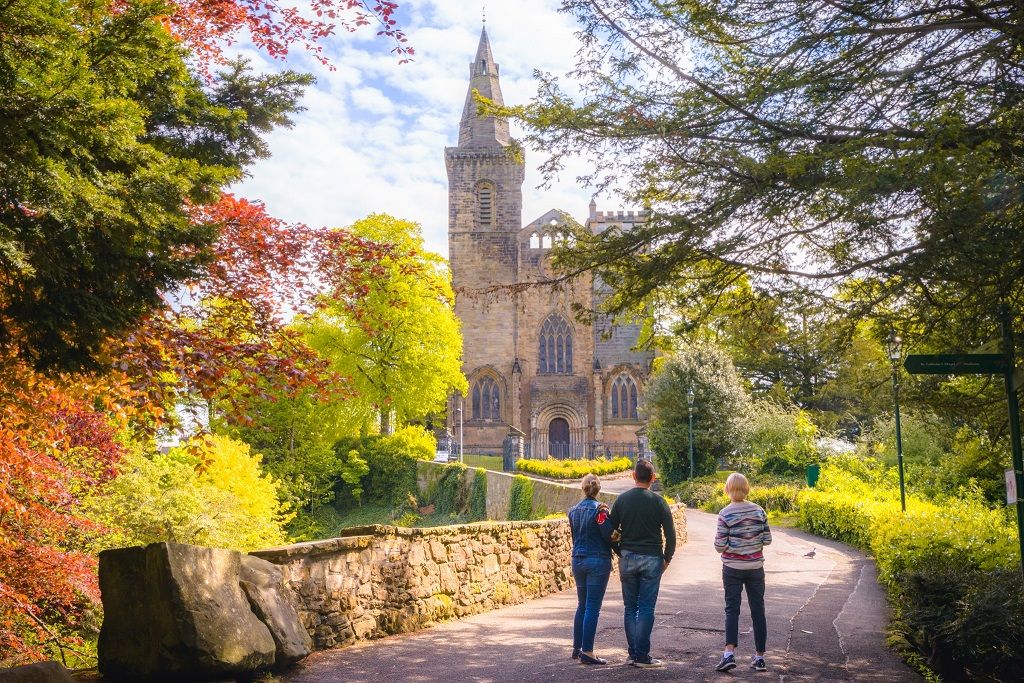 Three people approach Dunfermline Abbey amidst an autumnal landscape.