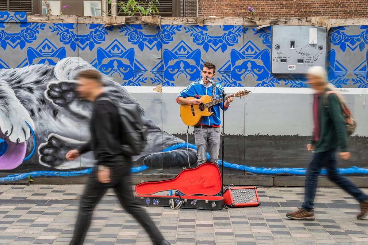 A person playing a guitar on the street with people walking by.