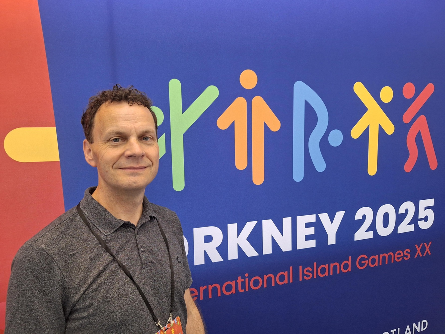 A man standing in front of a colourful sign that says Orkney 2025, International Island Games XX