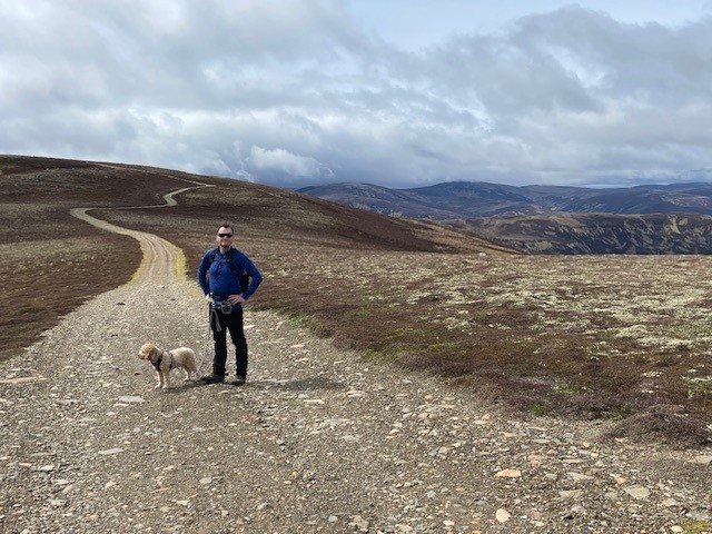 A man stands with his dog on a path in the middle of Cairngorms National Park. The path leads into mountains behind him.