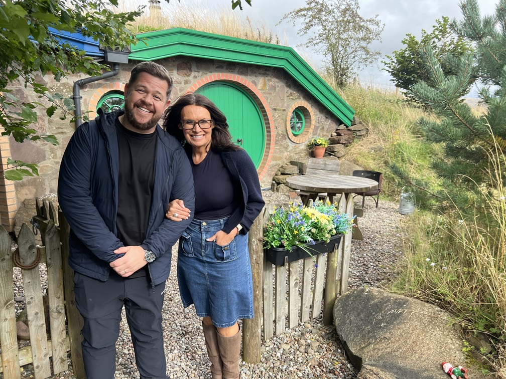 two people standing next to each other with a burrow, cottage style house in the background.