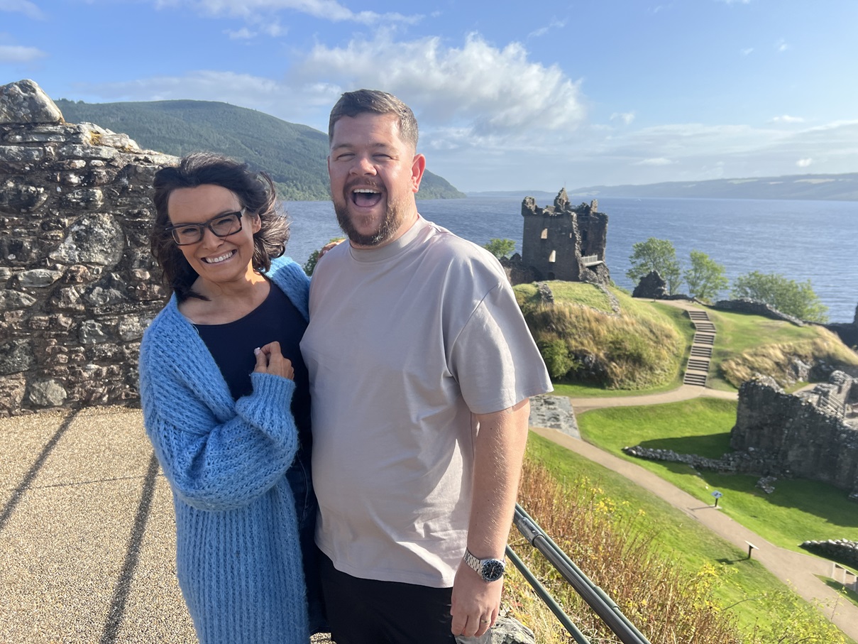 two people standing next to each other outside on a summers day with a picturesque backdrop of a castle ruin, and coast line.