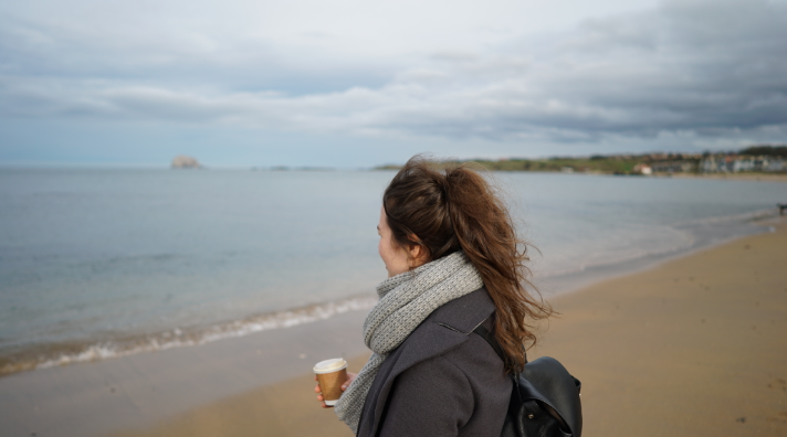 A girl with a scarf and a takeaway coffee stands on North Berwick beach