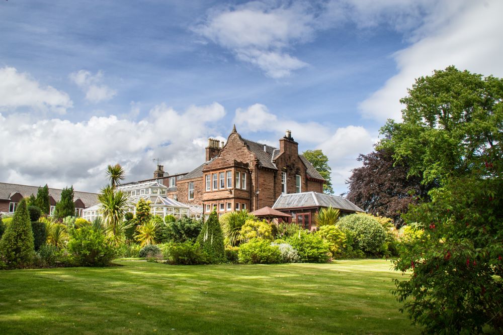 Image of exterior Auchrannie Resort behind a lavish garden with hedges lining a small pond and water feature.
