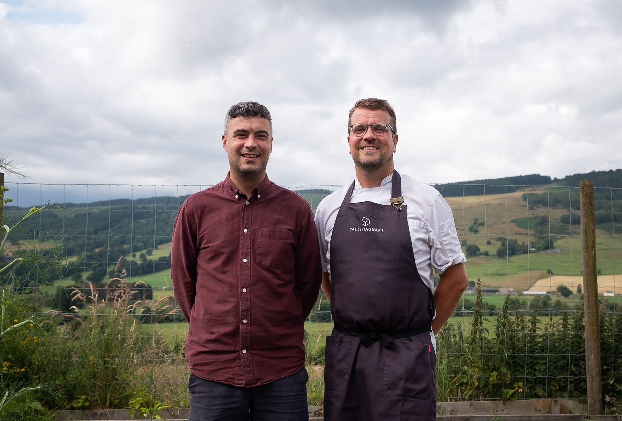 Two men standing in front of a field. One of them wears chef whites and an apron.