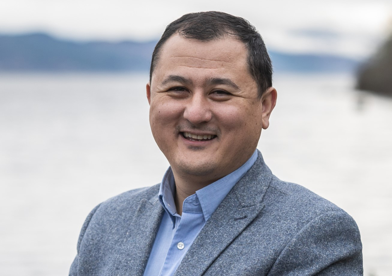 Image of a man, standing smiling to camera with a body of water behind him.