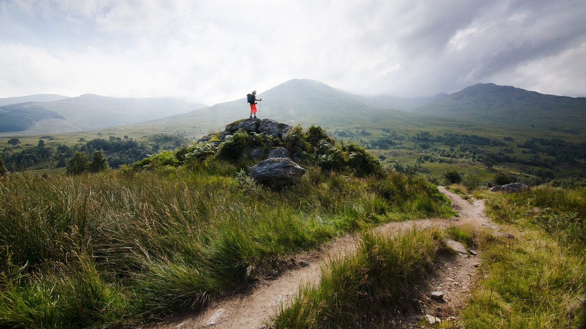A person standing on rocks, looking across to mountains while on the West Highland Way